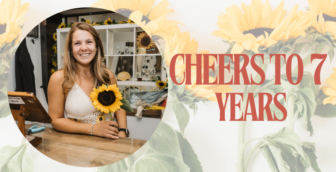 A digital birthday invitation template featuring a circular photo of a smiling woman holding a sunflower, set against a white background decorated with large sunflower graphics and the text 'CHEERS TO 7 YEARS' in red serif font.