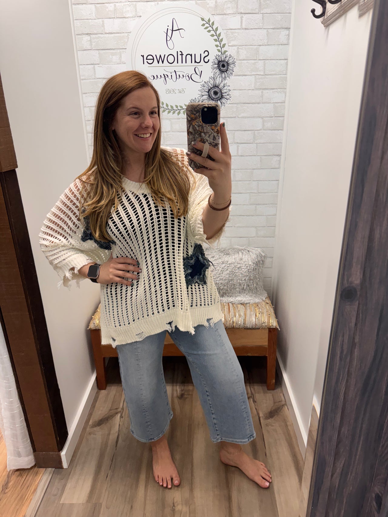 Woman taking a mirror selfie wearing a white sweater with blue stars and blue jeans in a fitting room.